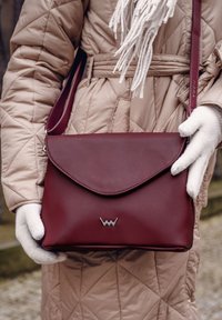 Burgundská kožená taška na ruku s klopovým uzávěrem, stříbrným logem a nastavitelným popruhem, držena rukama v rukavicích proti neutrálnímu pozadí.