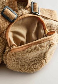 Tassengemaakt van een stof met een tan-textuur en een zachte buitenkant, met een ritsvak met een gladde beige voering en een metalen ritssluiting. Inclusief verstelbare banden.