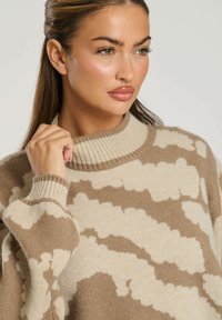 Beżowy sweter z dzianiny w brązowy wzór przypominający chmurki. Posiada ściągacz przy kołnierzu i mankietach, wykonany z miękkiego materiału.