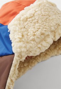 Chapeau coloré avec des sections orange, bleu et marron. Présente une doublure en polaire crème douce et texturée. Forme ronde avec des coutures visibles.