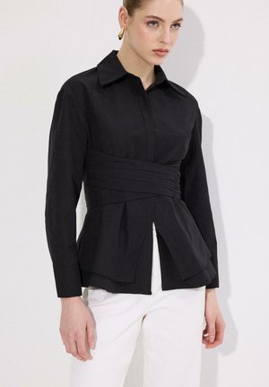 Femme portant un chemisier noir ajusté à manches longues avec un détail plissé à la taille et un pantalon blanc, debout devant un fond clair uni.