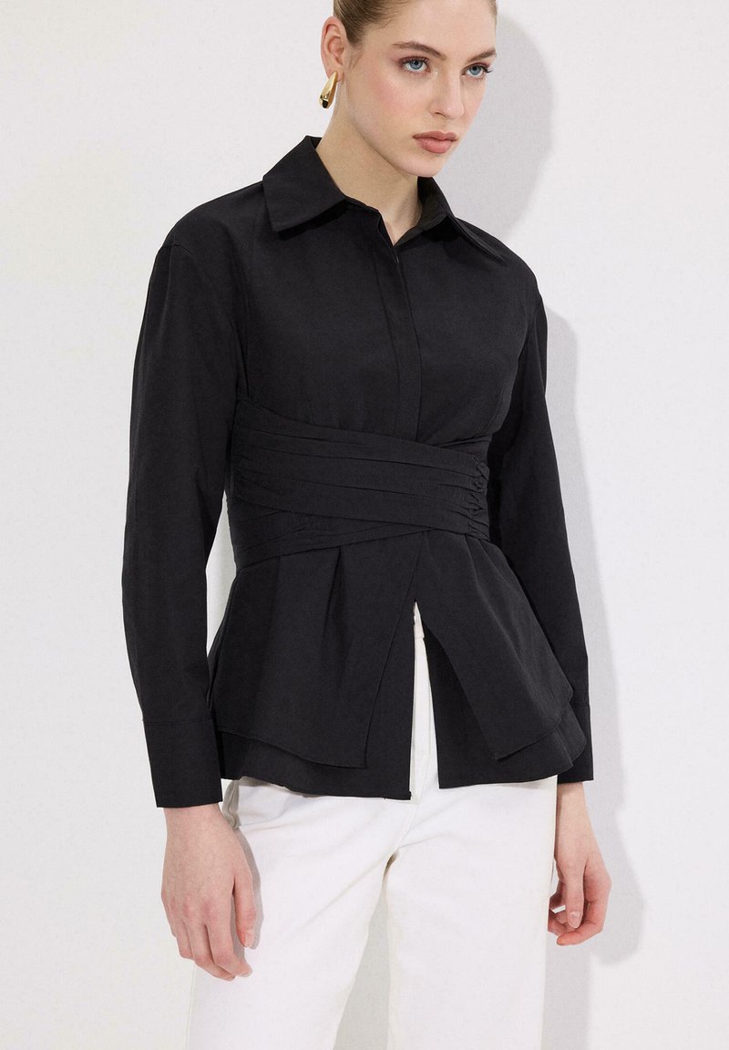 Γυναίκα που φοράει μαύρο εφαρμοστό μπλούζα με μακριά μανίκια και πλισέ λεπτομέρεια στη μέση, μαζί με άσπρο παντελόνι, στέκεται μπροστά σε απλό φωτεινό φόντο.