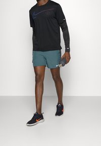 Svart Nike t-shirt med kort ärm och färgad logotyp, teal shorts och svarta skor med orange detaljer. Håller en grå vattenflaska.