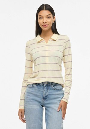 Žena s dlhými tmavými vlasmi v svetložltom košeľovom tričku s golierom a tenkými horizontálnymi pruhmi, svetlomodrých džínsoch, stojaca pred bielym pozadím.