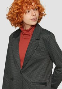 Szary kraciasty żakiet z klapami, pojedynczym zapięciem na guzik, dwoma przednimi kieszeniami, noszony na rdzawym golfie. Teksturowana tkanina.