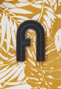 Logo en cuir noir en forme de "a" sur un tissu texturé avec un fond à motif floral blanc et jaune.