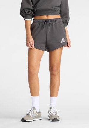 Frau trägt dunkelgraue New Balance Shorts mit Kordelzug, passendes cropped Sweatshirt, weiße Socken und beige New Balance Sneaker.