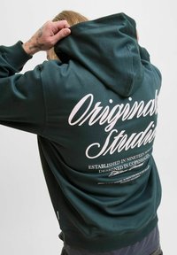 Hoodie vert foncé avec un grand logo en écriture blanche au dos, comportant un texte supplémentaire imprimé et une capuche avec cordon de serrage. Texture douce.