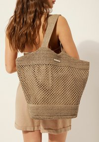 Bolsa de mano de punto en color beige con asas anchas, textura de tejido abierto, base sólida y etiqueta de marca visible en el lateral. Diseñada para uso casual.