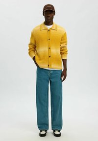 Geltonos gradientinės mezgimo kardiganas su apykakle, sagomis priekyje ir ribbintu apačios kraštu. Derinamas su laisvai krentančiomis mėlynomis džinsinėmis ir juodais batais.