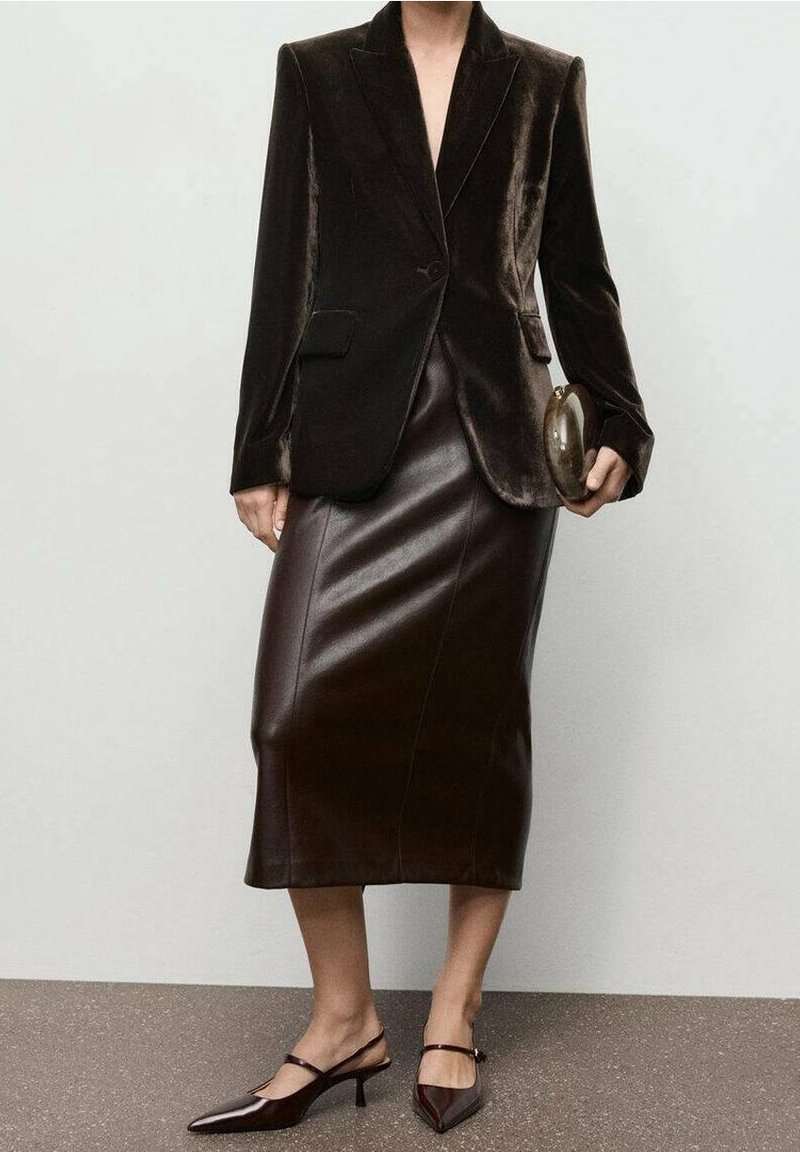 Femme portant un blazer en velours marron foncé, jupe midi en cuir assortie, escarpins marron foncé, tenant une petite pochette ovale.