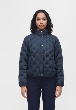 Femme à la peau moyenne et aux cheveux bouclés portant une veste matelassée bleu foncé avec des boutons dorés et un pantalon foncé, debout devant un fond uni.