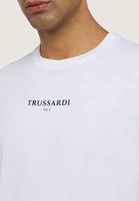 Camiseta de algodón blanca con cuello redondo, mangas cortas y la marca "TRUSSARDI 1911" impresa en negro en la parte delantera.