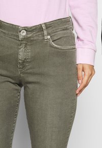 Olive zielone dżinsy o dopasowanym kroju, z kieszeniami z przodu i zamkiem błyskawicznym. Materiał ma delikatną teksturę, uzupełnioną srebrnymi detalami.