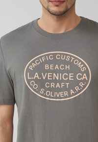 Graues Baumwoll-T-Shirt mit einem hellrosa ovalen Grafik und dem Text: "PACIFIC CUSTOMS BEACH L.A. VENICE CA CRAFT CO. S. OLIVER A.R.R."
