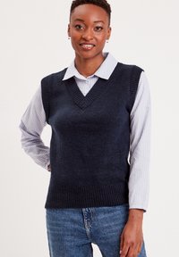 Mørkeblå strikket vest med V-udskæring og ribbet kant, båret over en stribet blå og hvid skjorte med krave, parret med blå denim jeans.
