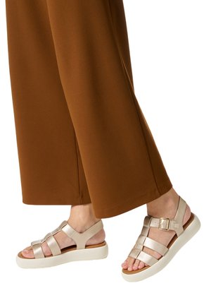 Piedi con sandali beige con cinturini, suole bianche e fibbie dorate, abbinati a pantaloni marroni a gamba larga.