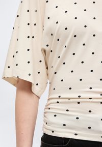 Μπεζ μπλούζα με μαύρες πουά βούλες, κοντά φαρδιά μανίκια και μαζεμένες πλευρές, φορεμένη με μαύρο παντελόνι.