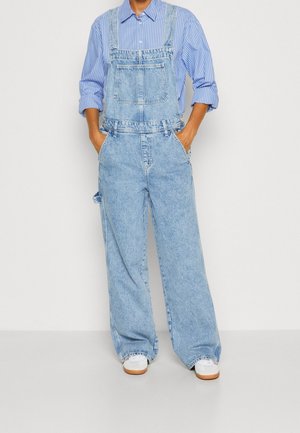 Personne portant une salopette en denim bleu clair à jambes larges, un chemisier rayé bleu et blanc aux manches retroussées, et des baskets blanches.