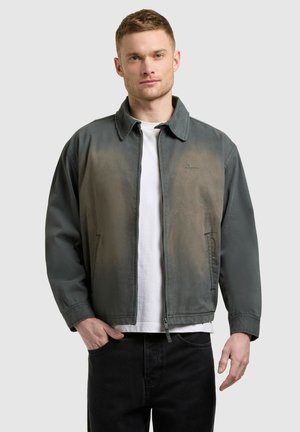 Ung mand iført falmet grøn lynlåstrøje over hvid t-shirt og sorte bukser, stående mod en ensartet lys baggrund.