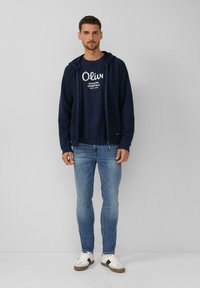 Donkerblauwe zip-up hoodie over een donkerblauw graphic t-shirt, lichtblauwe slim-fit jeans en witte sneakers met bruine accenten. Eenvoudige, casual outfit.