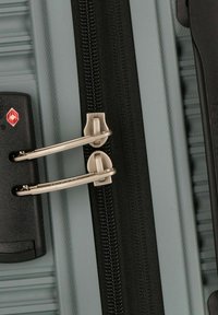 Valise rigide avec une finition gris clair, dotée d'une fermeture éclair noire et d'un cadenas argenté. Tirettes à double fermeture éclair notables pour une sécurité accrue.