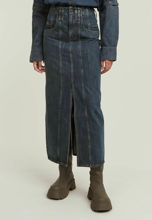 Jupe longue en denim foncé avec une fente à l'avant, portée avec des bottes épaisses foncées et une chemise en denim foncé assortie visible à la taille et aux manches.