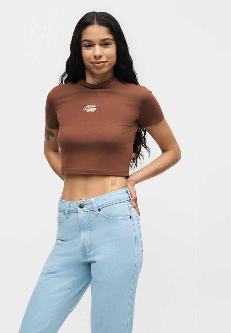 Frau mit langen dunklen Haaren, die ein braunes bauchfreies T-Shirt und hellblaue hoch taillierte Jeans trägt, steht mit einer Hand in der Hüfte.