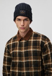 Szydełkowana granatowa czapka z naszywką. Brązowa koszula w kratę z sztruksowym kołnierzem i czarnymi guzikami, z pikowanym wzorem.