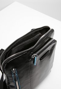 Bolso de cuero negro con múltiples compartimentos con cremallera, textura suave, acentos en cremallera azul contrastante y una correa para el hombro.