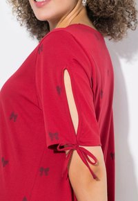 Chemise en coton rouge avec des manches courtes, dotée de fermetures à lien et d'ouvertures. Elle présente un motif de nœuds noirs sur le tissu.