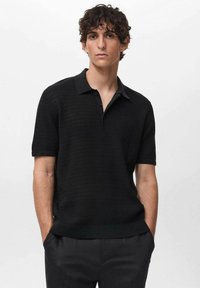 Polo shirt nero a maniche corte con design a maglia testurizzata, colletto piatto e patta con tre bottoni. Indossato con pantaloni su misura.