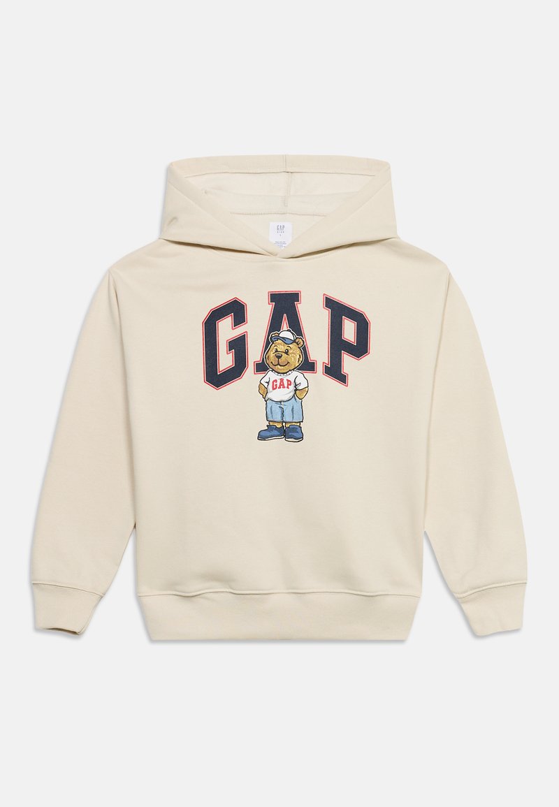 Sweat à capuche beige avec des lettres "GAP" bleu marine et rouge et un ours dessin animé portant un t-shirt GAP, un short bleu et une casquette blanche à l'avant.