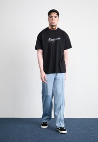 Zwarte katoenen t-shirt met wit "Carhartt" logo, gecombineerd met lichtblauwe broek en zwarte sneakers. Eenvoudige, relaxte pasvorm.