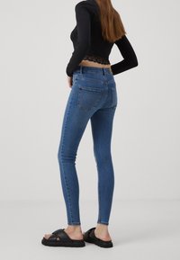Skinny jeans in blue denim, featuring a mid-rise waist, two back pockets, and a smooth texture. Paired with a black top and sandals.