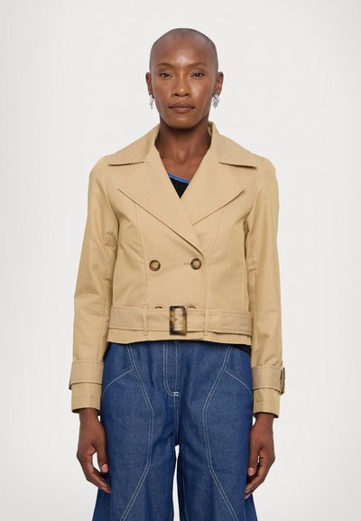 Veste beige coupée avec des boutons croisés, des revers larges et une ceinture. Présente une texture lisse et une silhouette carrée.