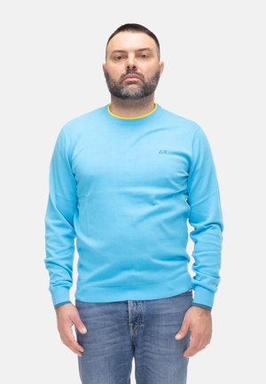Uomo con capelli corti e scuri e barba, indossa un maglione azzurro chiaro e jeans, in piedi davanti a uno sfondo grigio uniforme.