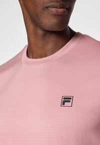 Lys rosa t-skjorte med korte ermer som har et teksturert mønster og en liten svart logo på brystet. Rund halslinje og mykt stoff.
