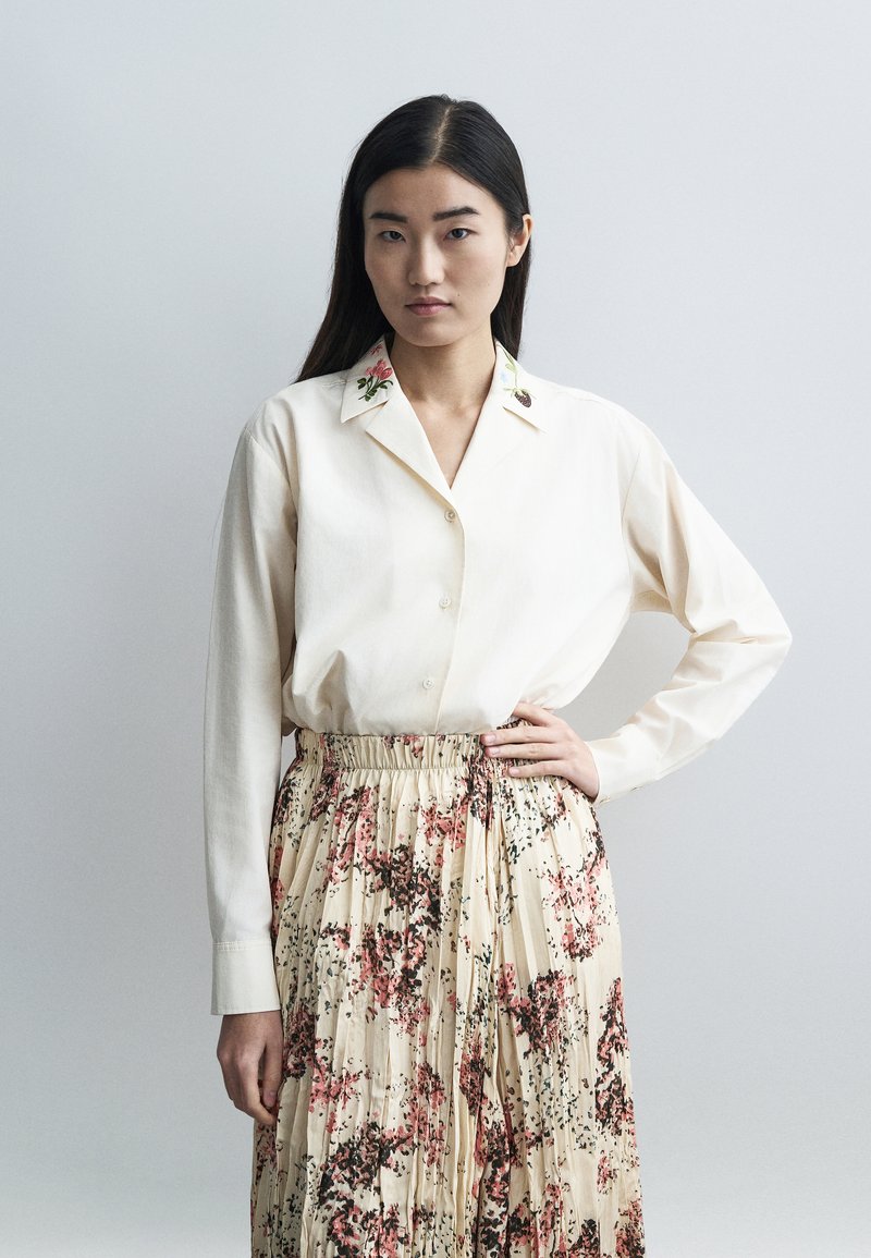 Mulher de cabelo longo e escuro veste blusa creme com gola bordada floral e saia creme plissada com estampa floral rosa e preta.