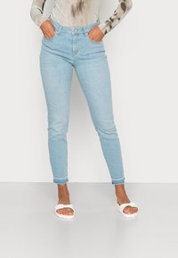 Jeans de ganga azul claro com um ajuste slim, apresentando um fecho de botão, bolsos frontais e punhos enrolados. Usados com sandálias brancas de slide.