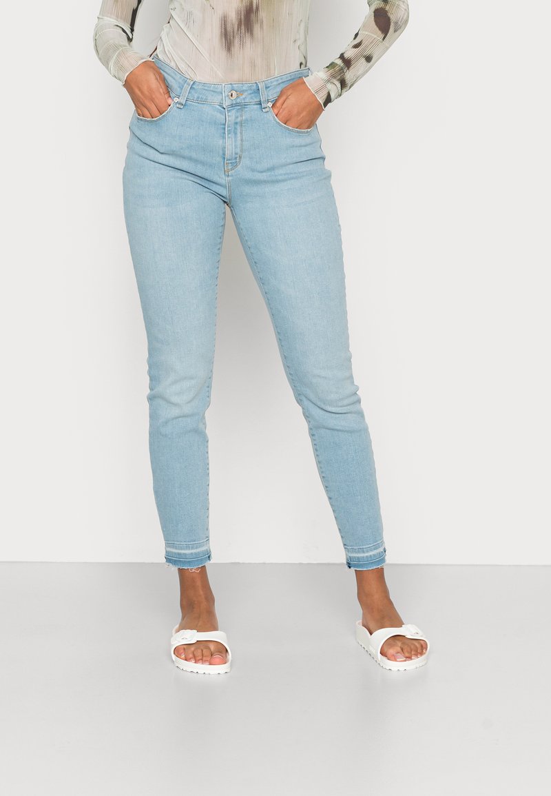 Jeans de ganga azul claro com um ajuste slim, apresentando um fecho de botão, bolsos frontais e punhos enrolados. Usados com sandálias brancas de slide.