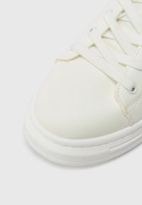 Hvit lær-sneaker med rundet tå, flate lisser og en teksturert gummisåle. Har søm langs kantene og minimal design.