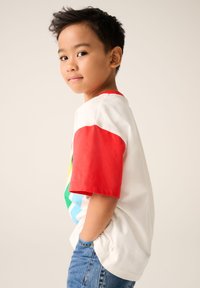 Vit t-shirt med röda ärmar som har ett färgglatt geometriskt mönster, tillsammans med blå jeans; enkel bakgrund.