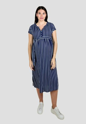 Těhotná žena stojící na jednobarevném pozadí, oblečená v modrobílých šatech s vertikálními pruhy po kolena a bílými teniskami.