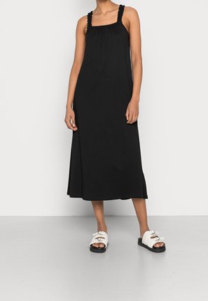Femme portant une robe midi noire sans manches avec des bretelles froncées et des sandales blanches à boucle, sur un fond uni.