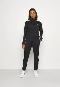 Czarny sportowy dres z białymi paskami po bokach, kurtka na zamek i zwężane spodnie. Wykonany z gładkiej tkaniny, o dopasowanym kroju. Białe trampki.