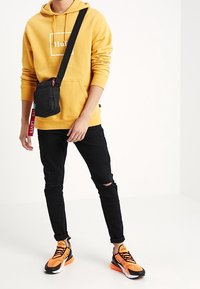 Felpa gialla con logo bianco, jeans neri strappati, sneakers arancioni e nere, e una piccola borsa a tracolla nera con dettagli rossi.