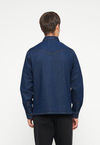 Giacca in denim blu scuro con maniche lunghe, polsini abbottonati e un design a yoke appuntito sul retro. Presenta dettagli di cucitura classici.