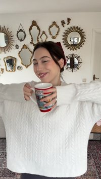 Roter Kaffeebecher mit grafischem Design, gehalten von zwei Händen, bekleidet mit einem grob gestrickten weißen Zopfpullover, vor einer Wand mit Spiegeln.