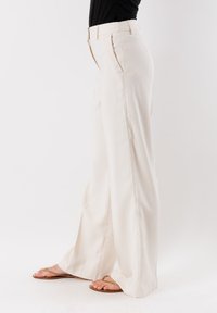 Pantaloni a gamba larga in tessuto beige chiaro con una texture liscia, caratterizzati da pince frontali e una vita strutturata. Abbinati a ciabatte beige.